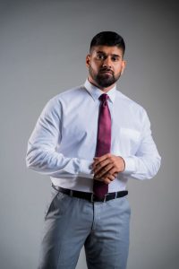 Professional man in a white shirt and maroon tie stands with hands clasped, looking at the camera.