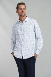 Man wearing a light blue patterned button-down shirt and navy trousers, standing with hands in pockets against a neutral backdrop.