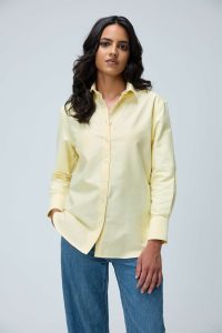 Woman wearing a pale yellow button-down shirt and blue jeans stands against a light gray background, neutral expression.