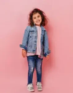 Smiling young girl with curly hair wearing a denim jacket, pink top, distressed jeans, and sneakers, standing against a pink backdrop.